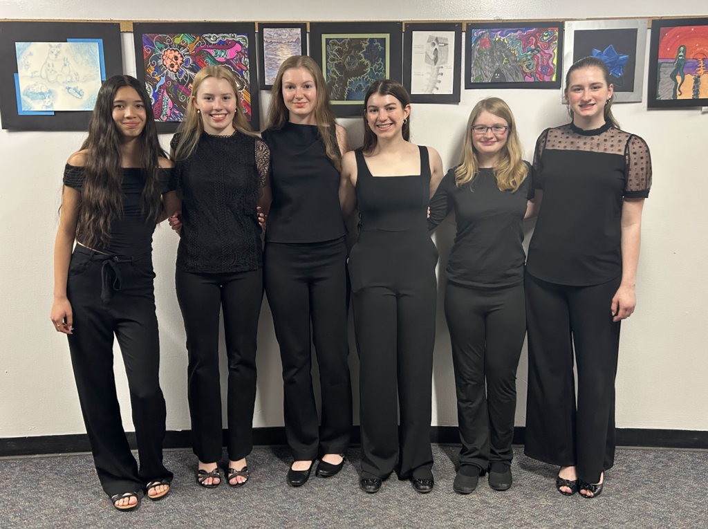 Six students dressed in black concert attire stand together smiling in front of a hallway art display featuring colorful student artwork after participating in the All State Music Festival.