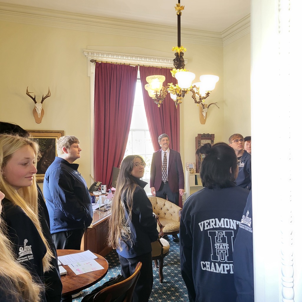 Members of the Fair Haven Union High School State Champion Bowling Team stand inside an office at the Vermont State House while listening to a guide. The room features tall red curtains, ornate lighting, antique furniture, and mounted deer antlers on the walls.
