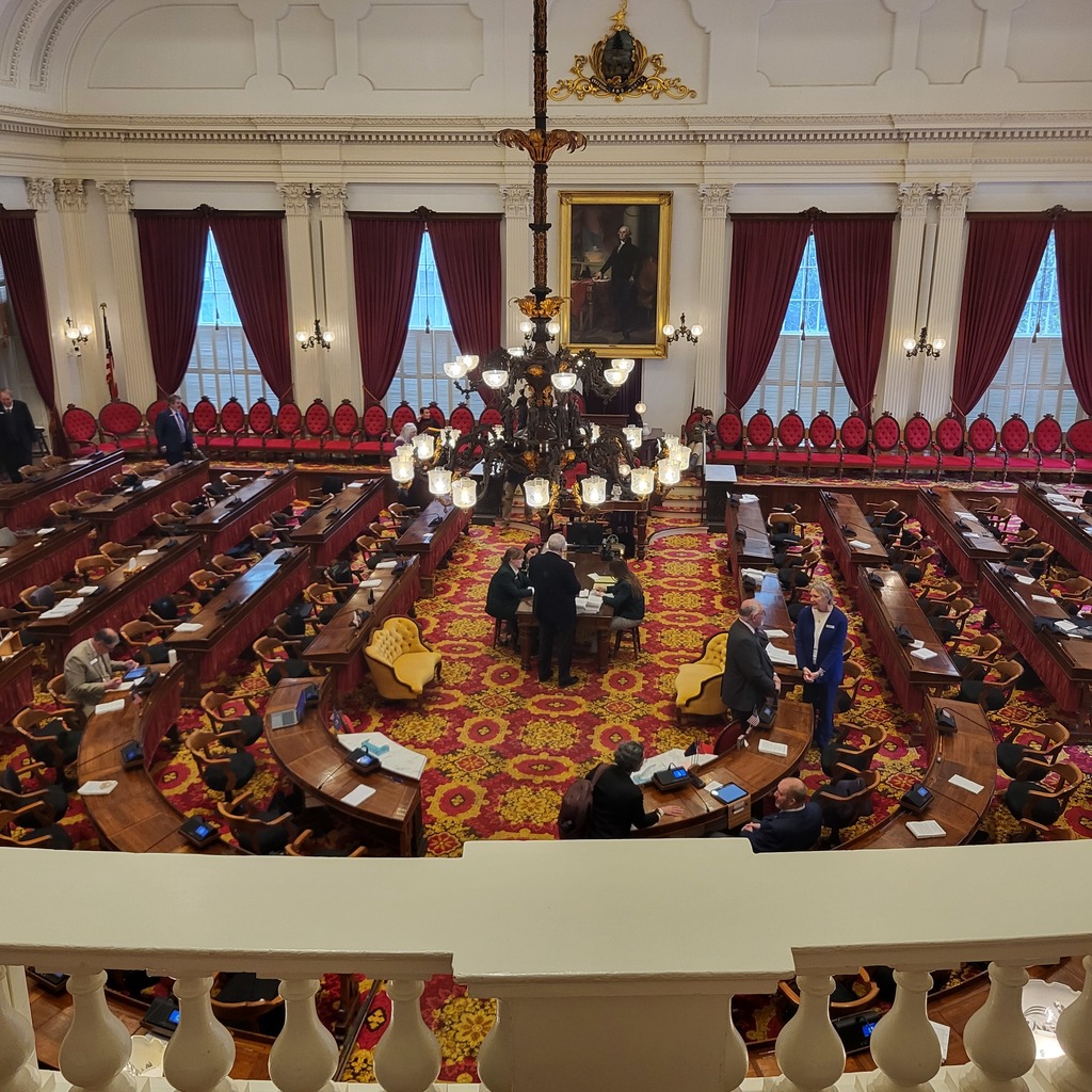 Wide view from an upper balcony overlooking the Vermont House chamber inside the State House. The chamber features rows of desks, red carpeting, tall white columns, large chandeliers, and portraits hanging on the walls while legislators and visitors gather below.