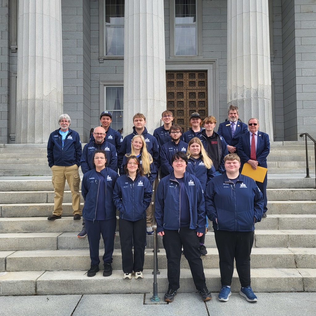 The Fair Haven Union High School State Champion Bowling Team poses for a group photo on the steps outside the Vermont State House with coaches and supporters. Students wear matching navy team jackets while standing in front of the building’s large stone columns and entrance doors.