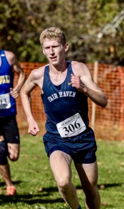 A Fair Haven Union High School athlete runs during a race while wearing a navy blue Fair Haven uniform and race bib number 306. Orange safety fencing and autumn trees line the course in the background as the runner strides forward during competition.