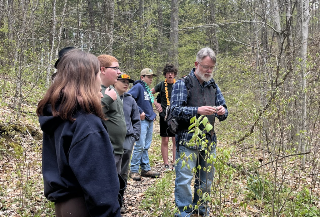Students stand along a wooded trail listening to an adult guide explain plants and invasive species during an outdoor educational activity at Half Moon State Park.