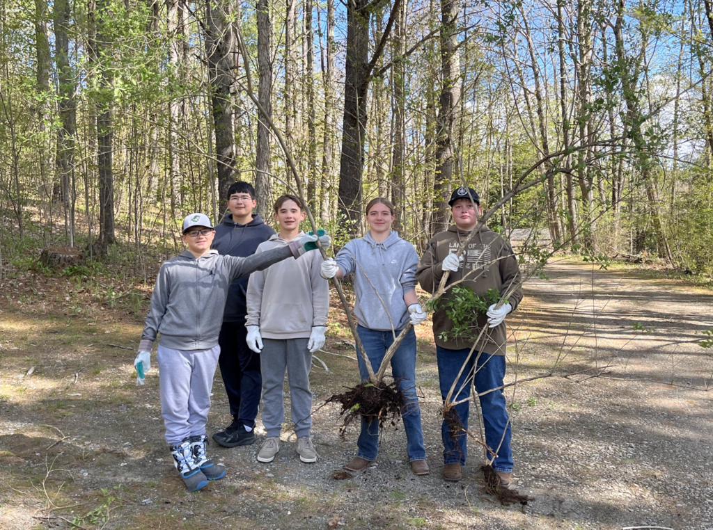 Four students wearing gloves stand together outdoors holding uprooted invasive plants during a volunteer cleanup project at Half Moon State Park.