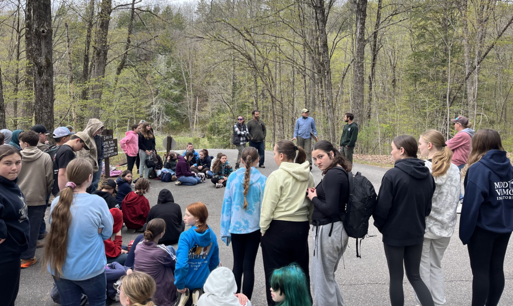 A large group of middle school students gathers on a park roadway at Half Moon State Park while listening to adults give instructions before beginning volunteer work in the wooded area.