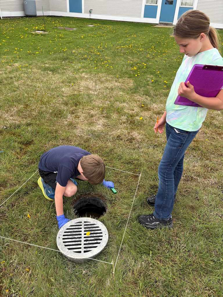  A young boy in blue gloves kneels inside a square string plot on a lawn, looking into an open drainage hole. A girl stands nearby holding a purple clipboard, observing the study area. 