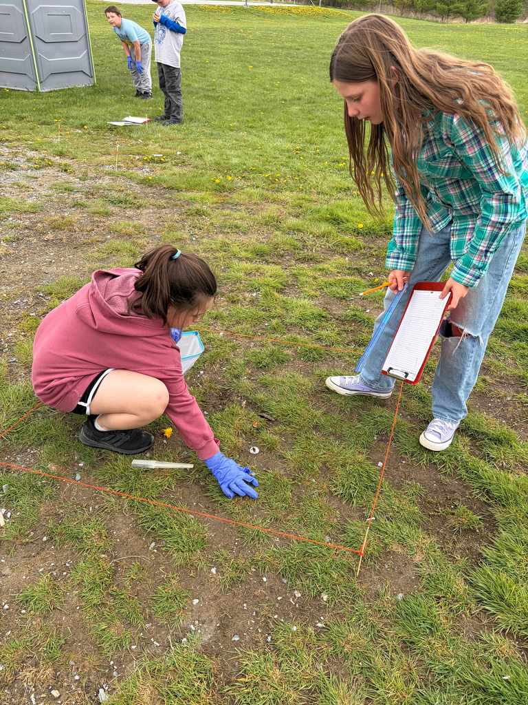  A girl in a green plaid shirt holds a red clipboard and a ruler, standing over a square string plot on a patchy lawn. Another girl in a pink hoodie kneels inside the plot, reaching toward the ground with blue gloved hands.
