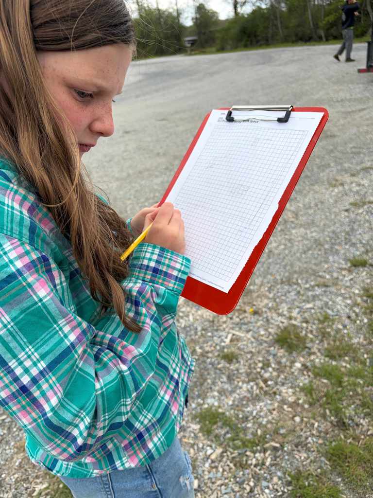  A close-up of a girl with freckles and long brown hair wearing a green plaid shirt. She is focused on writing or drawing on a grid-lined "Problem String Work Space" sheet attached to a red clipboard.