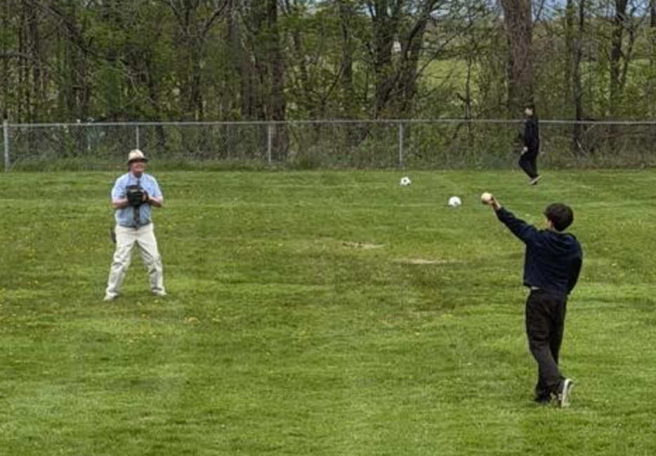 Students and an adult play catch on a grassy field outdoors near a chain-link fence and trees. One student is throwing a baseball while another person wearing a glove prepares to catch it.