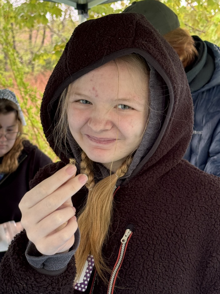 A close-up of a smiling young girl wearing a dark, fuzzy hooded jacket with her blonde hair in braids. She is holding up a small, dark archaeological fragment between her thumb and forefinger for the camera to see.