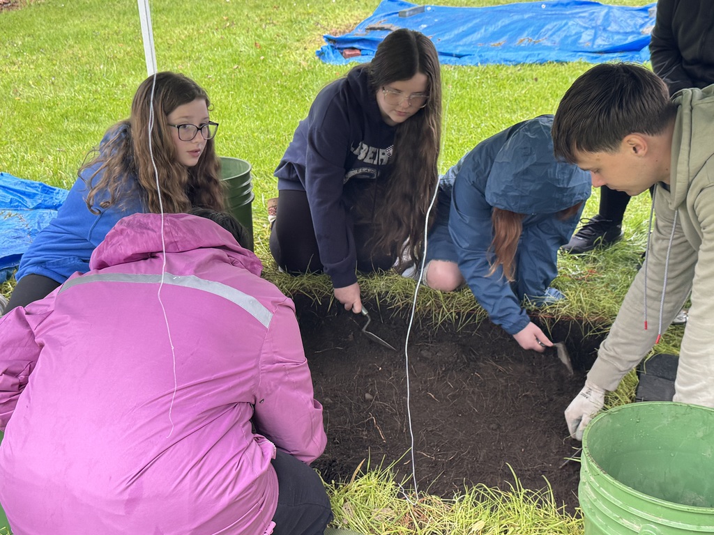 Four students are kneeling on a grassy lawn around a small, square excavation unit in the dirt. They are using hand trowels to carefully dig and scrape the soil. A green bucket sits nearby, and a blue tarp and a pink rain jacket are visible in the frame.