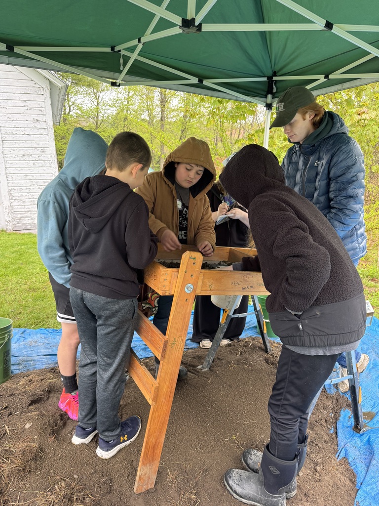 A group of five students stands under a green pop-up tent, working at a large wooden sifting screen. They are spread out around the frame, using their hands to pick through dark soil and rocks to find artifacts. An old white farmhouse with weathered siding is visible in the background. .