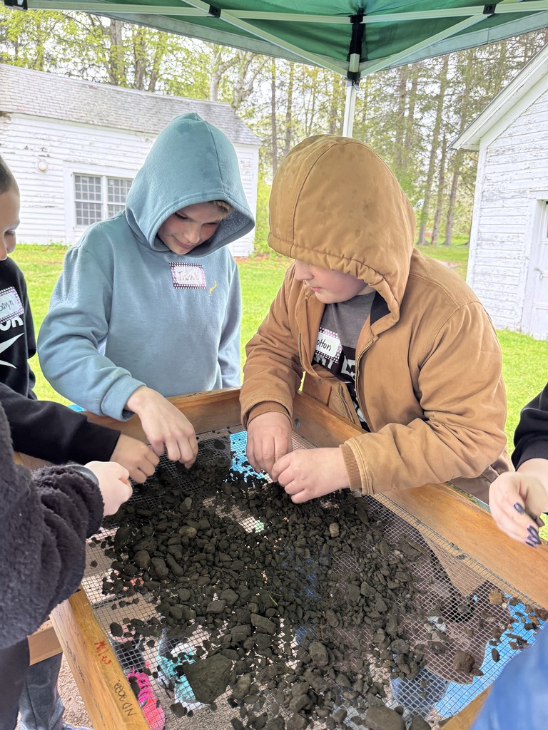 A medium shot focusing on two students at the sifting station. One student in a light blue hoodie and another in a tan Carhartt-style jacket are leaning over the wire mesh screen, intently searching through a layer of dark clumps of earth and stones.