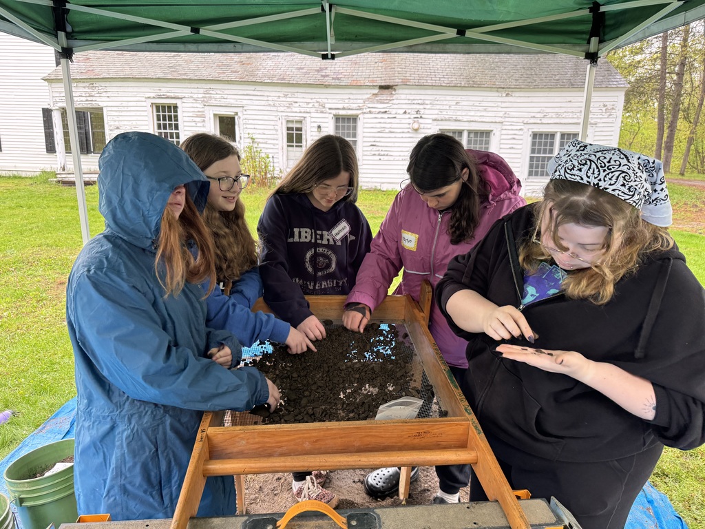 A group of five students stands under a green pop-up tent, working at a large wooden sifting screen. They are spread out around the frame, using their hands to pick through dark soil and rocks tundo find artifacts. An old white farmhouse with weathered siding is visible in the backgro. IMG_4375.jpg A wide shot showing several students gathered under a green canopy tent. In the center, a wooden shaker screen is mounted on a frame, and students are actively sifting through a pile of dirt. A large blue tarp is spread on the ground beneath the station to catch the processed soil.