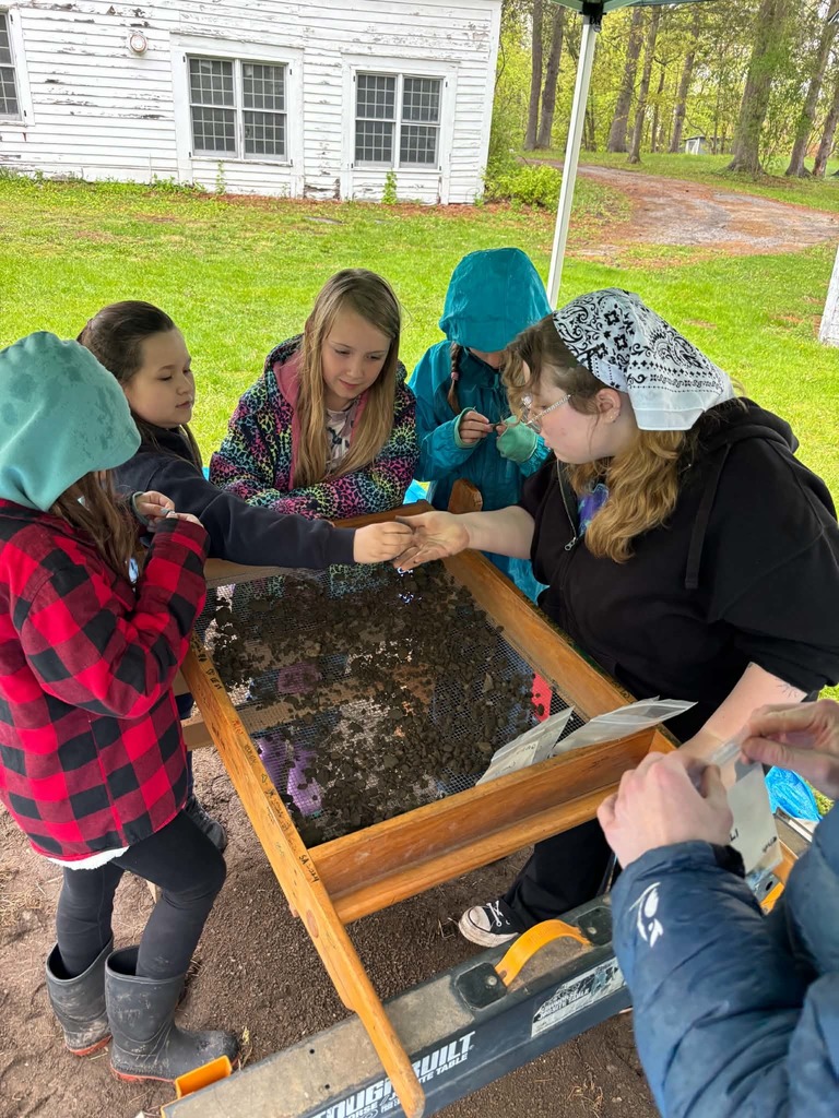 Archaeological Sifting Under a white canopy, a group of young students and an instructor work with a large wooden archaeological sifting screen. The screen is filled with dark soil, and the students are carefully picking through the dirt to find small artifacts, placing them into plastic bags.