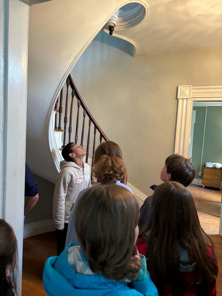 Indoor Architectural Tour A group of students stands in a hallway of an old house, looking up in awe at a unique architectural feature. In the center, a white spiral staircase curves upward toward a circular skylight in the ceiling, which casts soft light into the room.