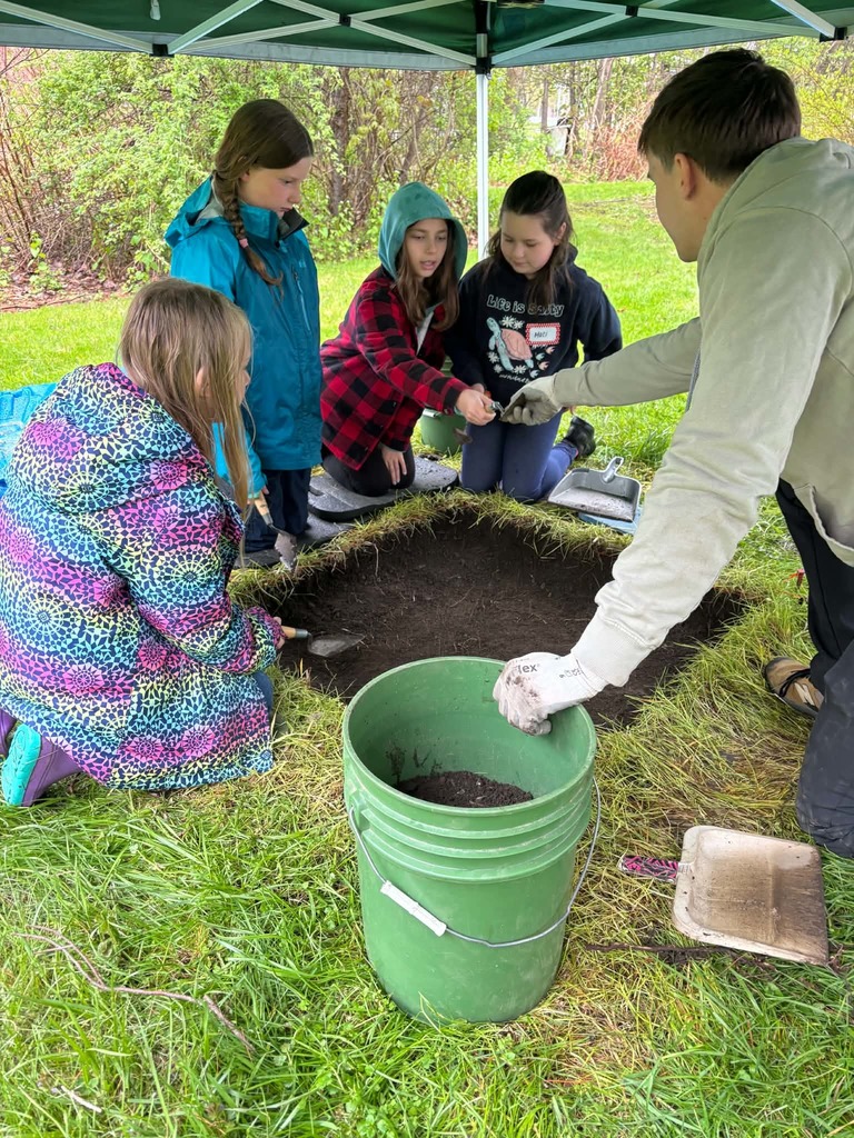 Active Excavation Site A group of children kneels around a square, excavated pit in the grass under a green tent. They are using small hand trowels to carefully dig into the dark earth. An instructor in a tan hoodie and work gloves assists them, while a green bucket nearby holds the removed soil.