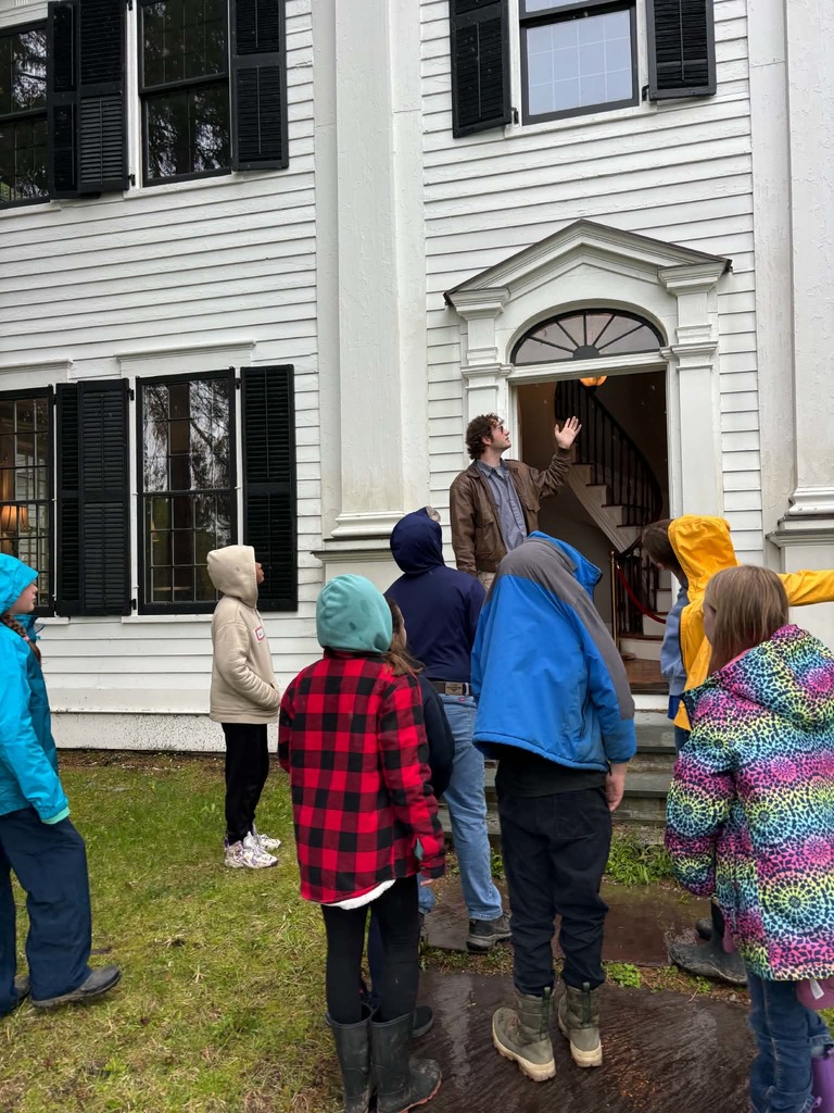Historic House Entrance A tour guide in a brown leather jacket stands in the arched doorway of a historic two-story white house with black shutters. He gestures toward the interior as a group of students, mostly in rain gear and hoods, looks on. A glimpse of the white spiral staircase is visible through the open door.