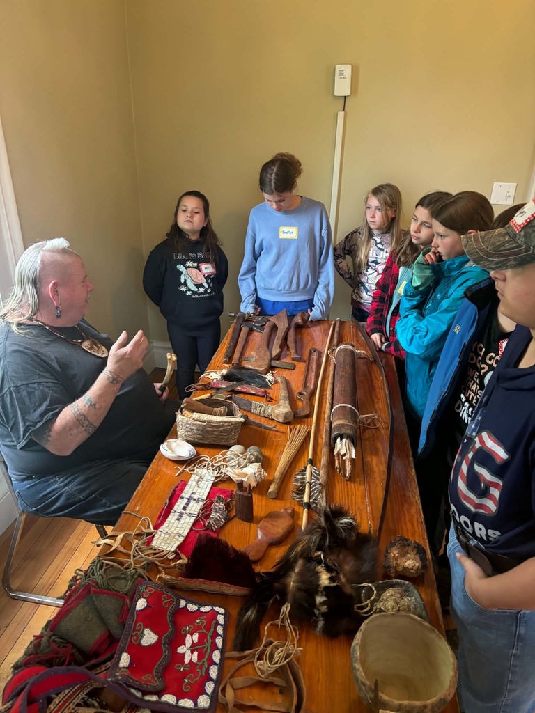Indigenous History Presentation An indoor shot of a group of children gathered around a long wooden table. An instructor with a white mohawk and arm tattoos is seated at the head of the table, gesturing as he speaks. The table is covered in Indigenous artifacts, including hand-carved wooden tools, a bow and arrows, woven baskets, beaded textiles, and animal furs.