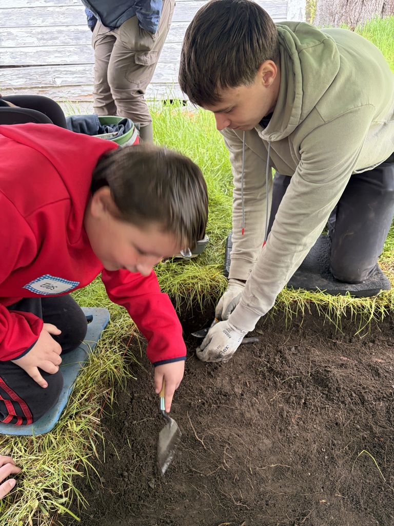 A young boy in a bright red hoodie and an older teen in a tan hoodie kneel on padded mats next to an archaeological test pit. They are carefully using small hand trowels to scrape away layers of dark soil in a squared-off section of grass.