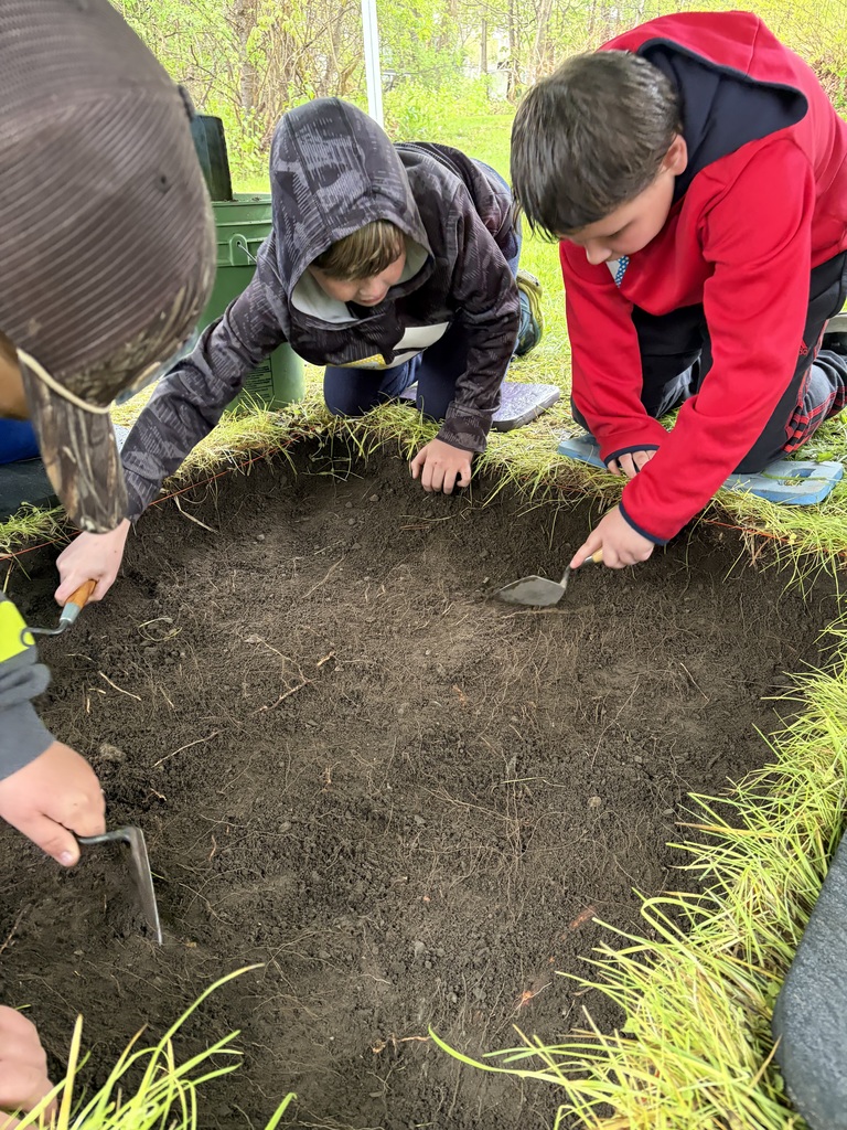 : A young boy in a bright red hoodie and an older teen in a tan hoodie kneel on padded mats next to an archaeological test pit. They are carefully using small hand trowels to scrape away layers of dark soil in a squared-off section of grass.