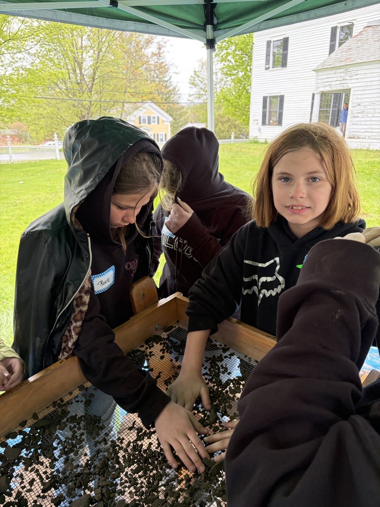 Under a green canopy, three students lean over a wooden sifting screen. They are using their hands to push dark soil and small rocks through the mesh to search for hidden artifacts.
