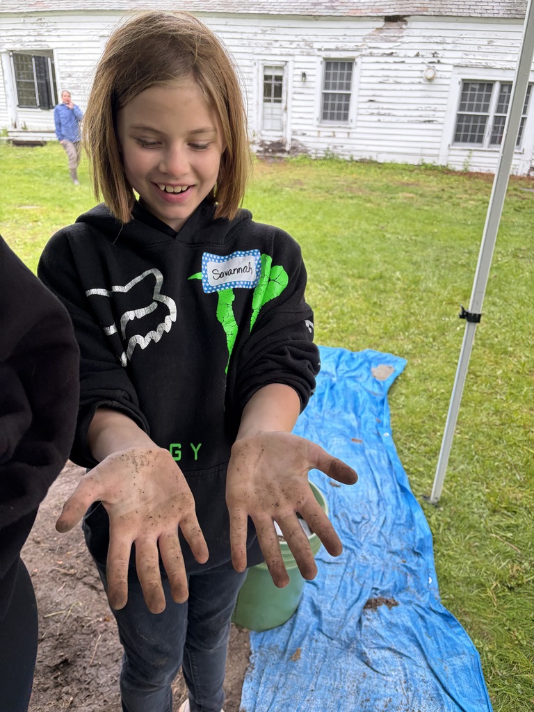 A student wearing a black Fox Racing hoodie, smiles as she holds her dirt-covered palms out toward the camera. In the background, a white farmhouse with peeling paint sits behind a blue tarp on the lawn.