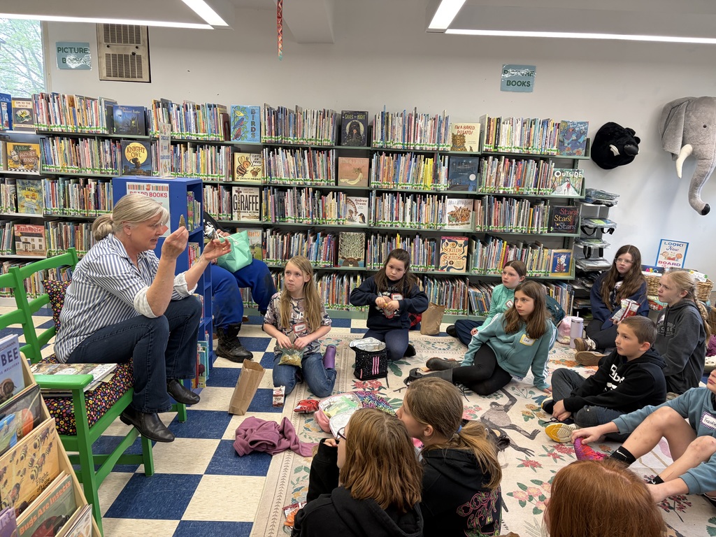 Inside a library filled with colorful bookshelves, an instructor sits on a green chair and holds up a small artifact for a group of students. The children are sitting on a patterned rug, listening intently during a lesson about their findings.