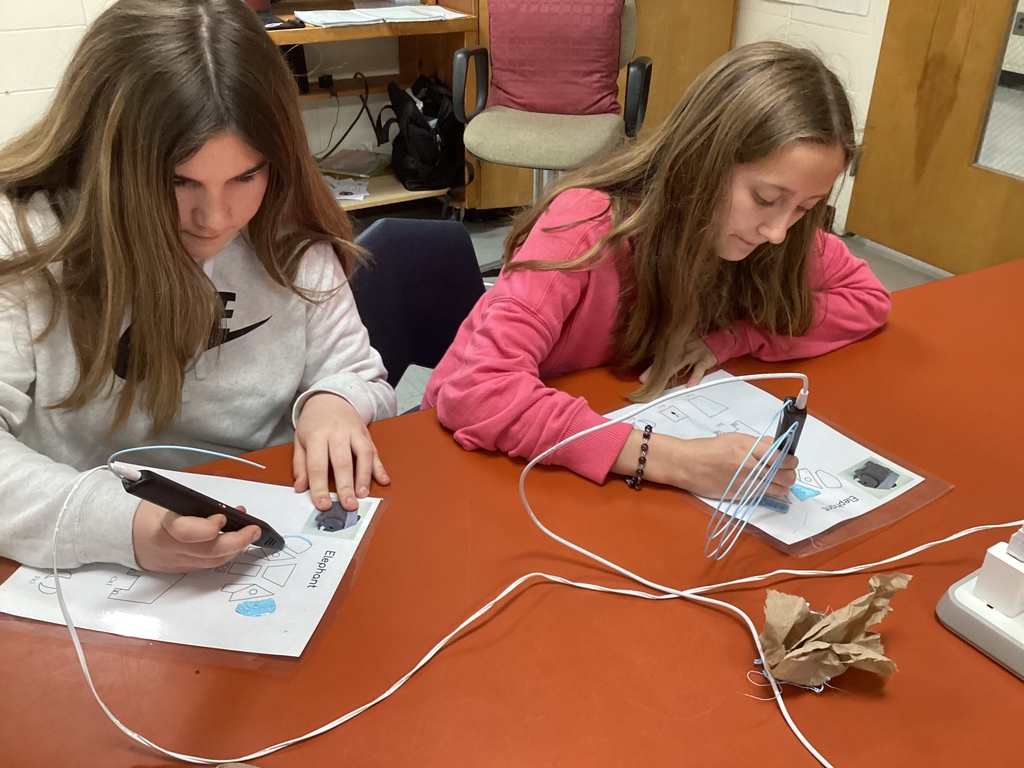 Two students sit side by side at a library table using 3D pens to create colorful designs on worksheets during a hands-on library activity.