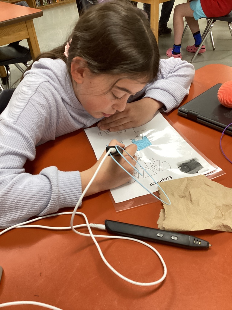 Student wearing a light purple sweater carefully uses a black 3D pen to trace shapes on a worksheet while seated at a library table.