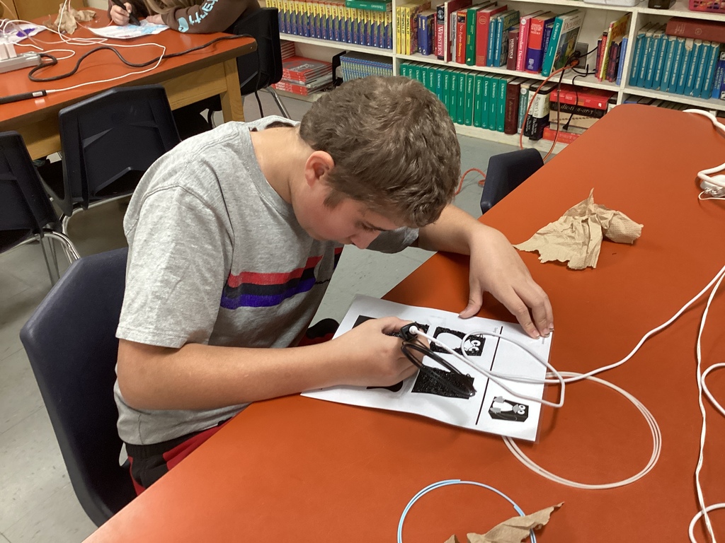 Student in a gray T-shirt carefully uses a 3D pen to trace a black design on a worksheet at a classroom table.