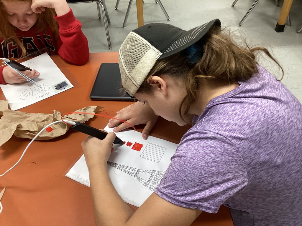 Student in a purple shirt and baseball cap leans over a worksheet while using a 3D pen to create red shapes and patterns.