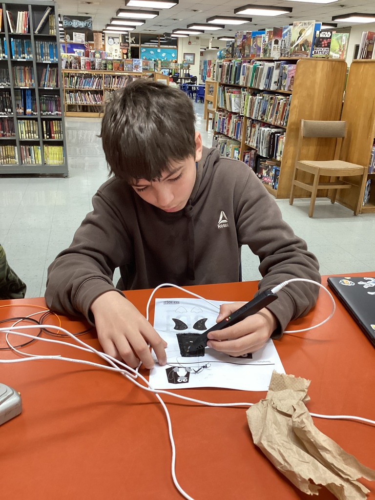Student in a brown hoodie concentrates while using a 3D pen to create a black design on a worksheet in the school library.