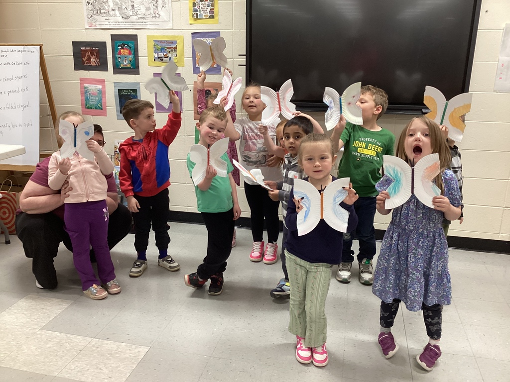 A group of Pre-K students stand together in a classroom proudly holding colorful paper plate butterflies they created after reading Waiting for Wings in library class. The students smile and pose in front of a classroom display and large screen while showing off their butterfly crafts.