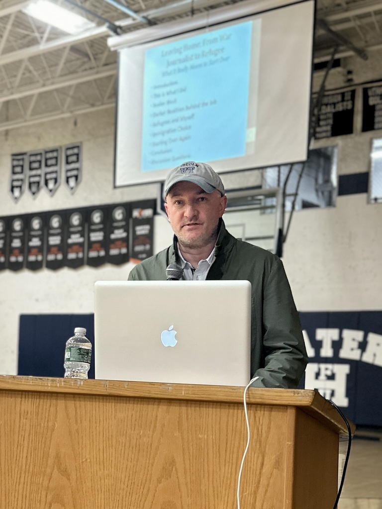 A guest speaker stands behind a podium with a laptop and microphone in a school gymnasium while giving a presentation. A projection screen with presentation text is visible in the background.
