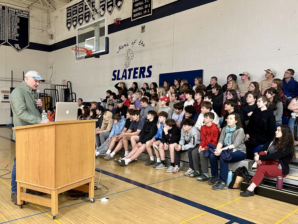 Photojournalist Dmitry Beliakov speaks to a large group of middle school students seated on gym bleachers at Fair Haven Union High School. Students and staff listen attentively during the presentation.
