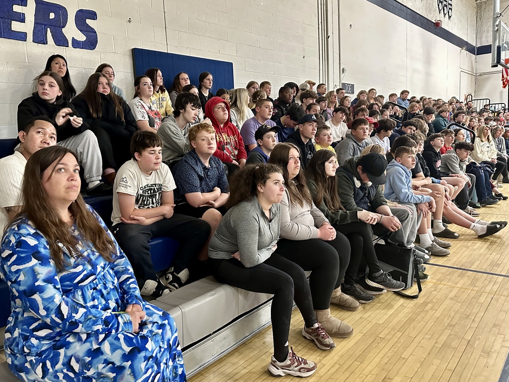 Middle school students and staff sit closely together on gym bleachers, listening to a guest speaker during an assembly presentation in the school gymnasium.