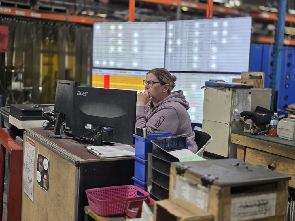 An employee sits at a workstation inside the manufacturing facility, monitoring production information on multiple computer screens surrounded by office and factory equipment.