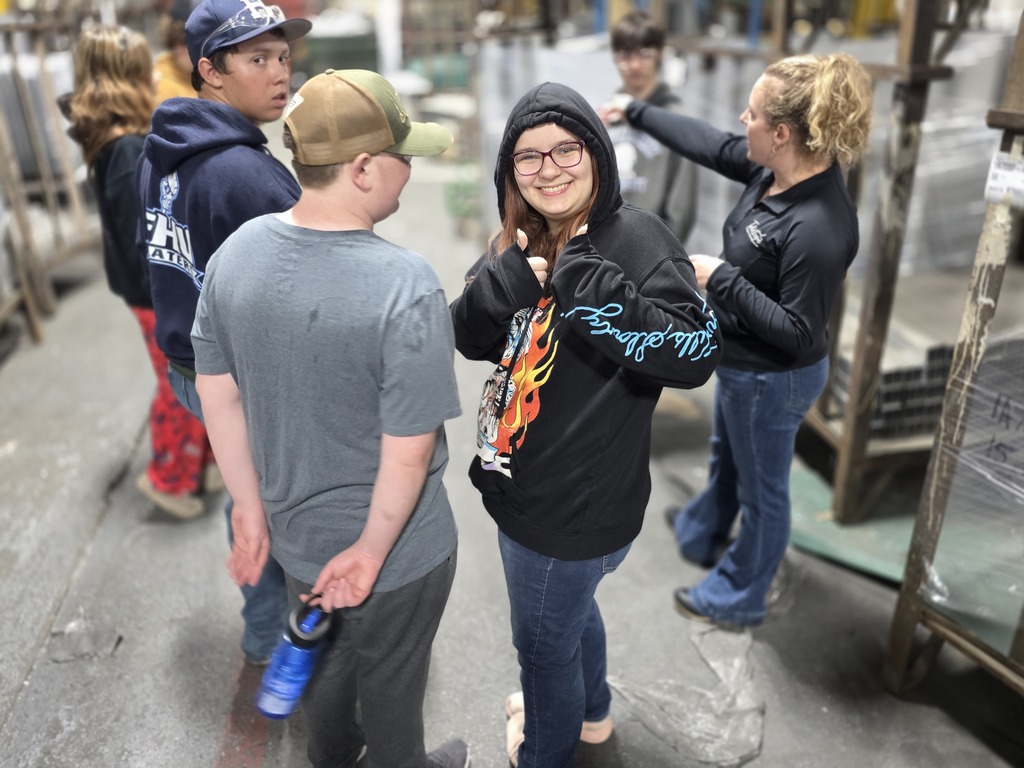 Several students stand inside the Telescope Casual manufacturing facility while listening to a staff member explain equipment and production processes. One student in a black hoodie smiles and gives a thumbs up toward the camera.