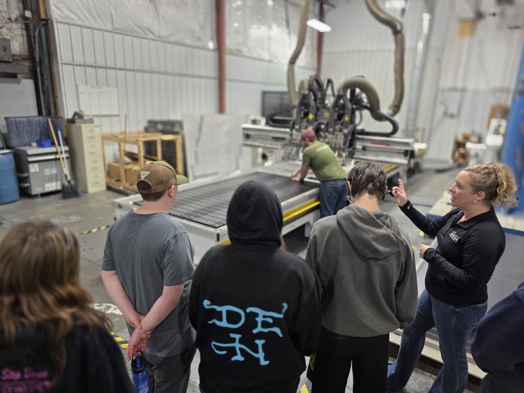 Students gather around a large industrial cutting machine while a staff member explains how the equipment is used during the manufacturing process.