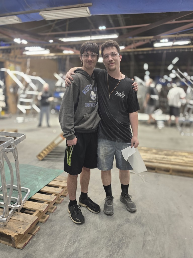 Two students stand together smiling inside the Telescope Casual production facility with manufacturing equipment and partially assembled furniture frames visible in the background.
