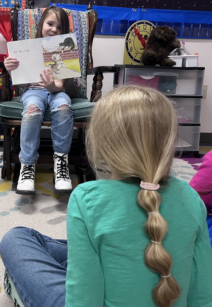  Close-up of Reading Time Alt Text: A young girl with long brown hair sits in a wooden rocking chair, holding open a children's book titled "Taco sees a cat." She is wearing ripped blue jeans and white combat boots. In the foreground, the back of another child's head is visible, showing blonde hair styled in a long bubble ponytail secured with several hair ties. In the background, a yellow toy clock and a stuffed brown animal sit on a plastic storage bin.  