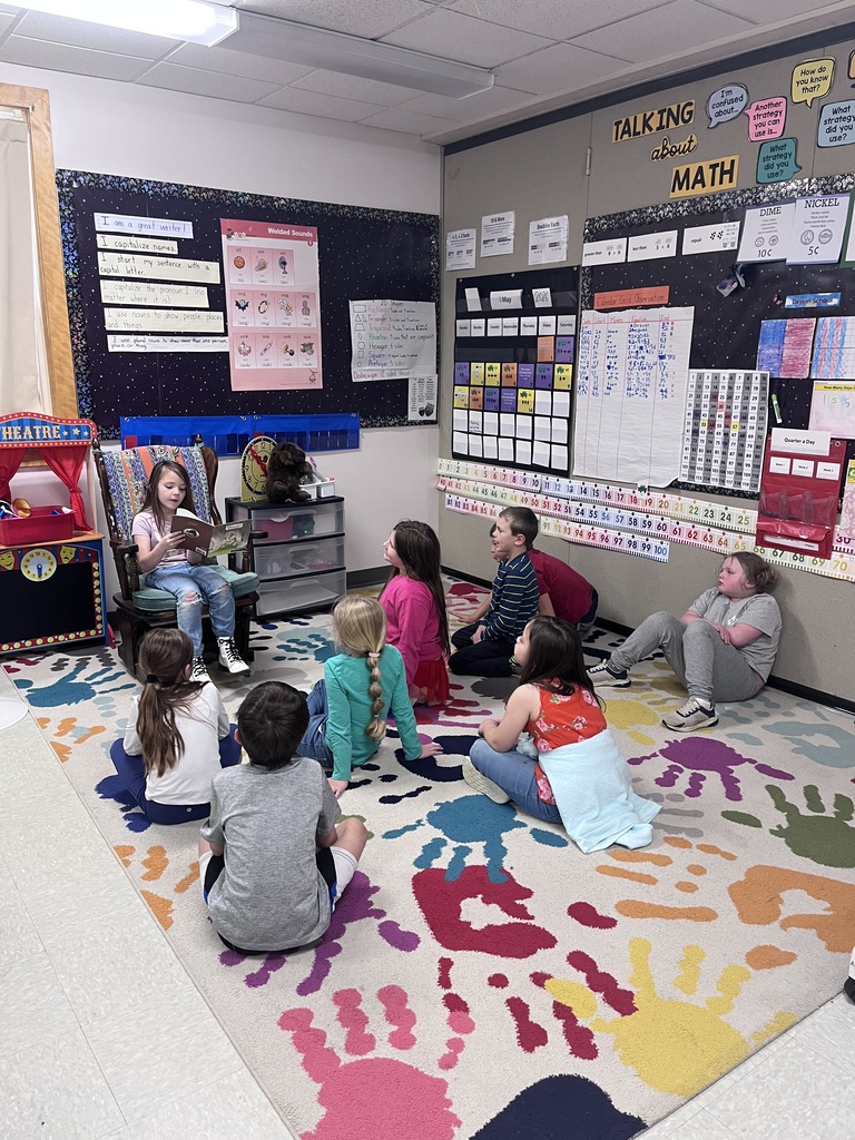 Wide View of the Classroom Alt Text: A classroom scene where a young girl sits in a rocking chair and reads aloud to a group of six children sitting on a colorful rug decorated with large, multi-colored handprints. The room is filled with educational posters, including a "Talking about Math" board, a calendar, and a "Welded Sounds" chart. The walls are decorated with dark blue fabric and various learning aids, and a small toy "Theatre" set stands to the left of the reader.