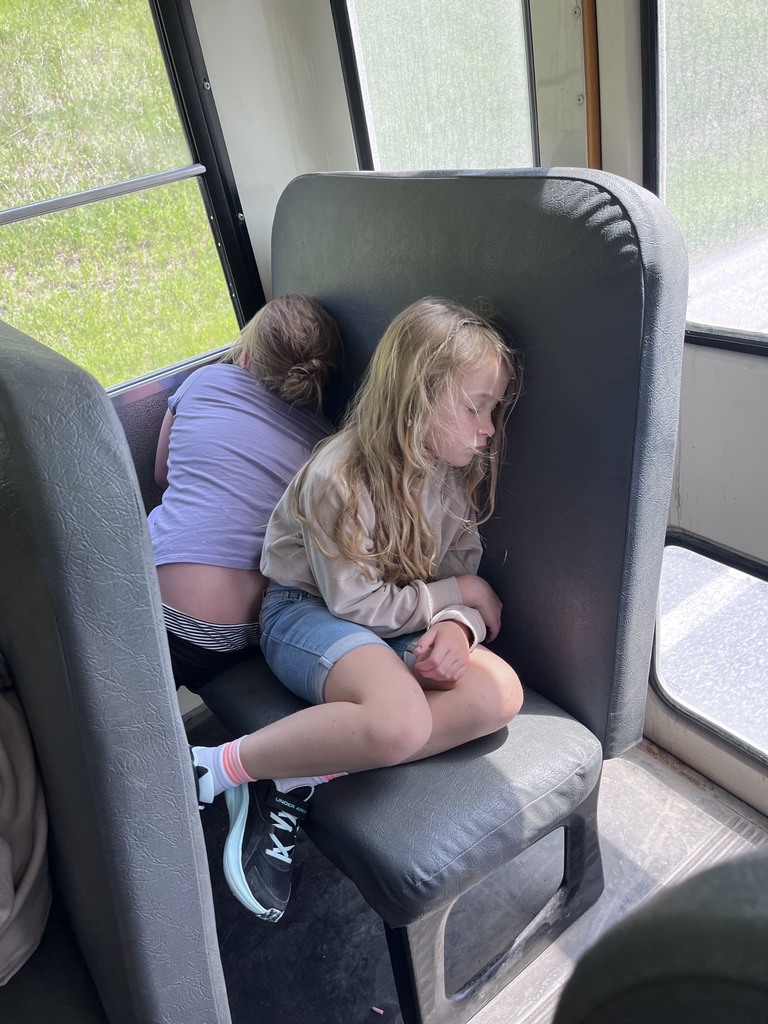 Two young girls napping on a gray school bus seat. The girl in the foreground has long blonde hair and is curled up with her eyes closed, wearing a beige sweatshirt and denim shorts. The girl behind her is leaning her head against the window panel with her back to the camera.