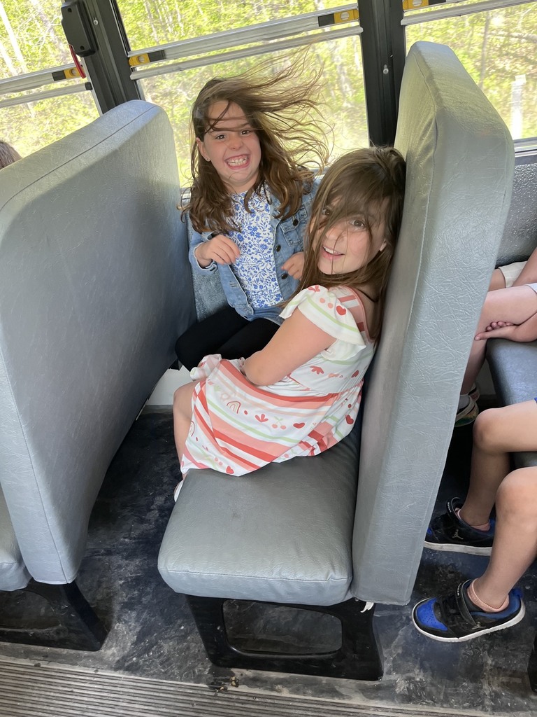 Two young girls smiling and laughing on a school bus seat. The girl in the back wears a denim jacket, and her long hair is blowing wildly in the wind from an open window. The girl in front wears a white dress with pink and orange stripes and looks back toward the camera with a smile.