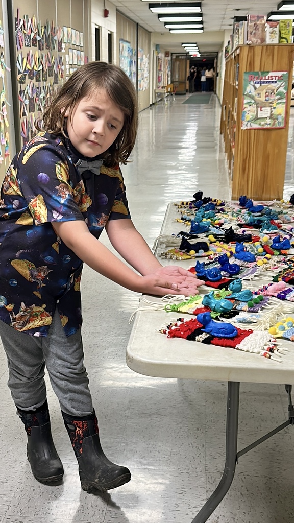 Alt text: A student wearing a patterned shirt and bow tie gestures toward a table filled with colorful student art projects displayed in a school hallway.