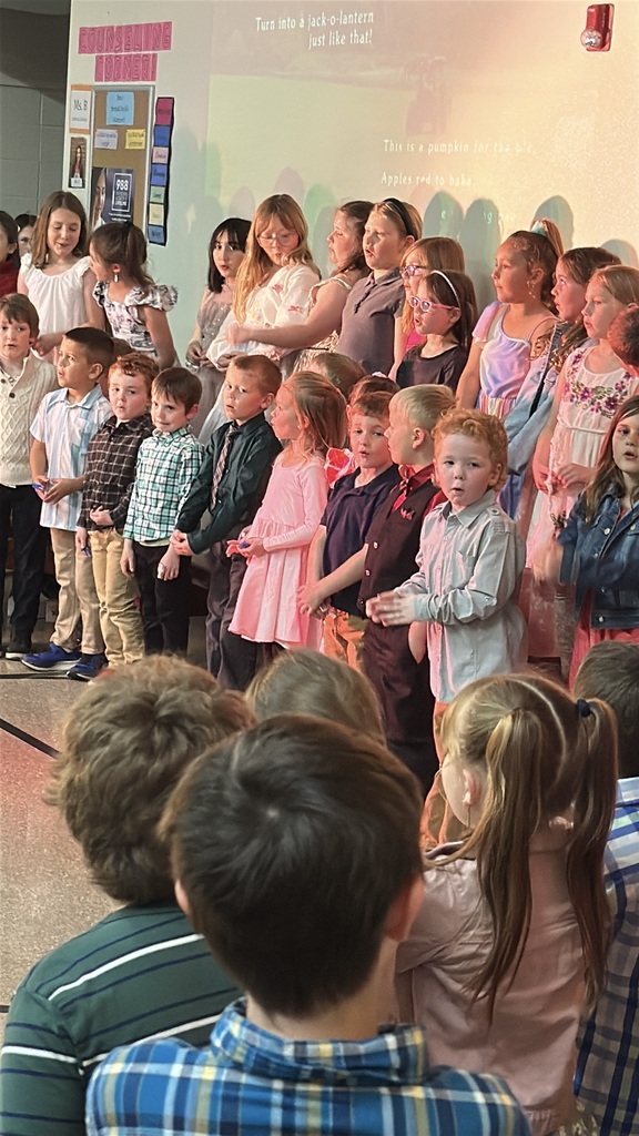 Elementary students stand together performing during a school concert while classmates and families watch from the audience. A projected image and song lyrics appear on the wall behind them.