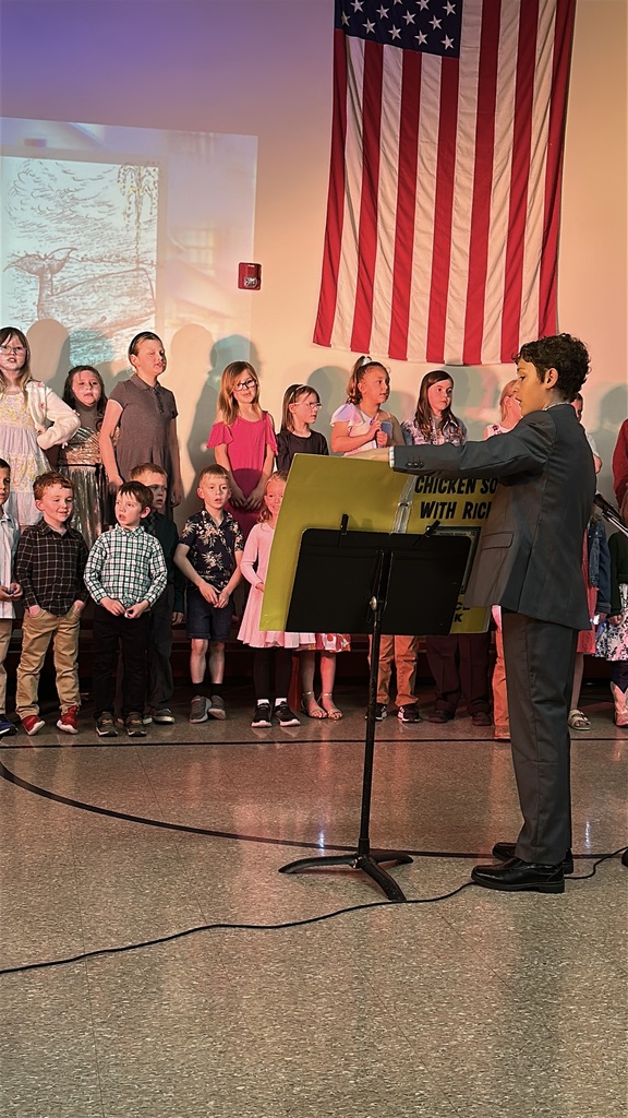 A student conductor in a suit leads a group of elementary students during a school performance beneath a large American flag hanging on the wall.