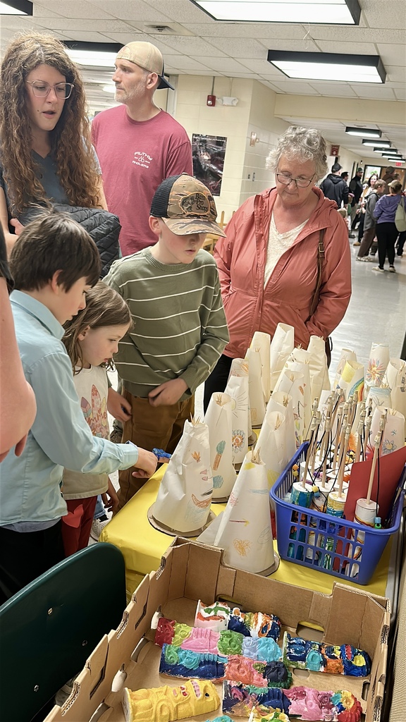 Alt text: Families and students gather around a table filled with student-created art projects and colorful paper sculptures during a school showcase event.