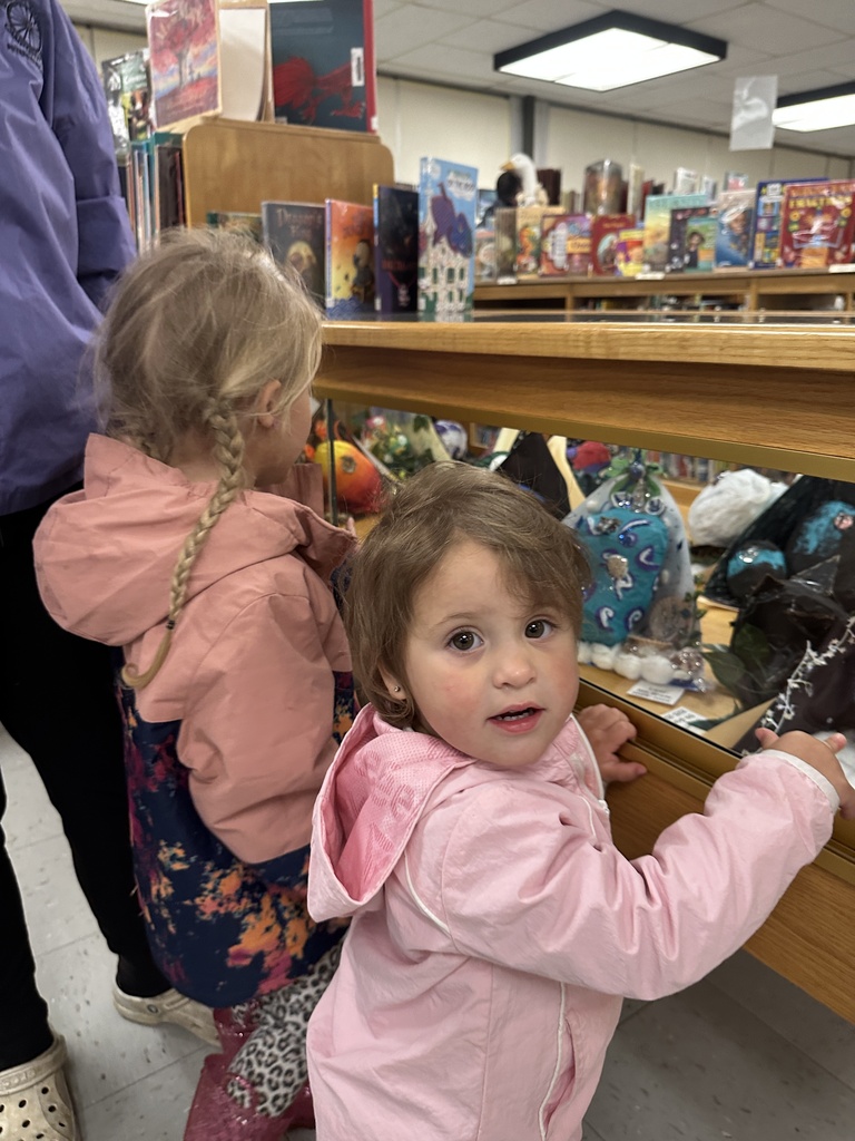 Alt text: Two young children in pink jackets look at student artwork and decorative pieces displayed inside a library display case.