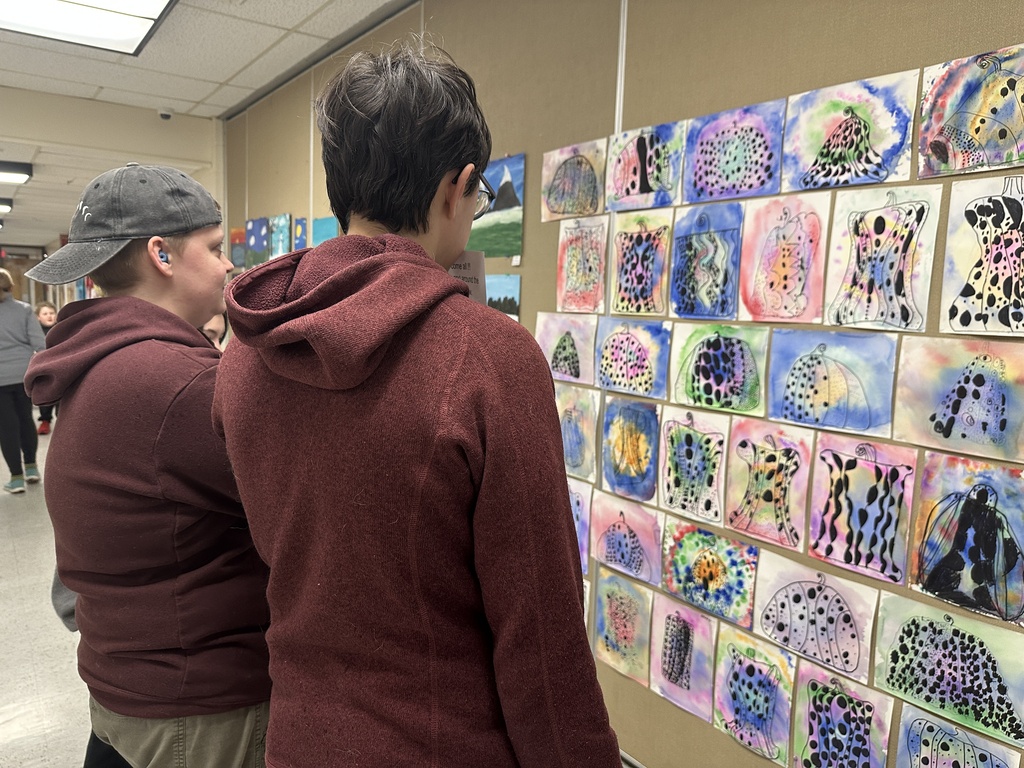 Alt text: Two people stand in a school hallway viewing a colorful wall display of student artwork featuring abstract and patterned designs.